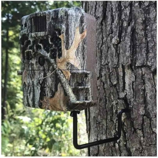 Camouflage trail camera mounted on a tree with a metal L-bracket in a forest setting.